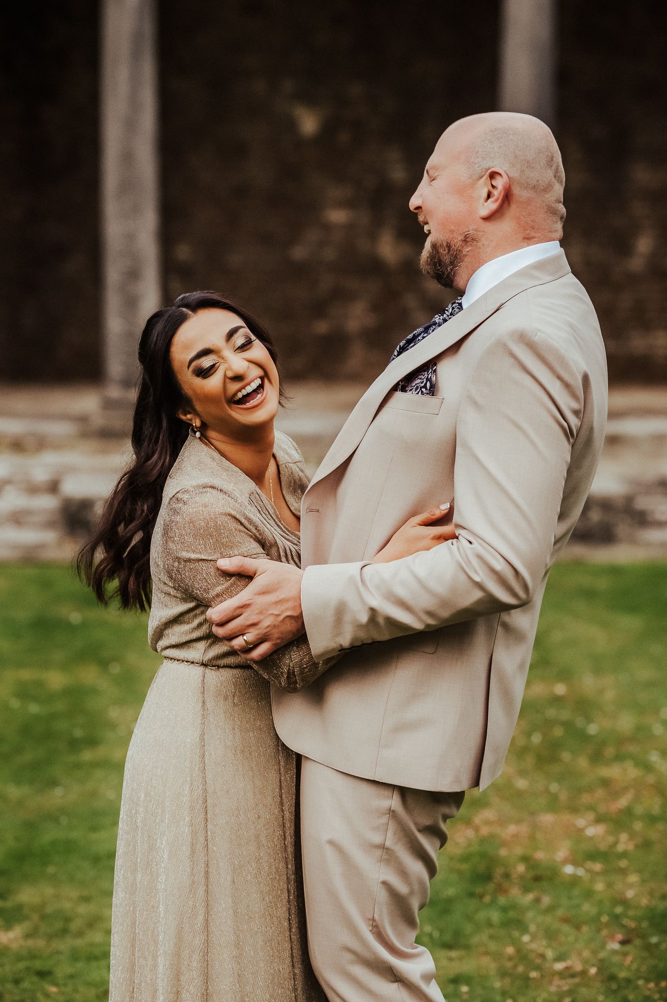 Two people embracing and laughing together outdoors wearing matching beige formal attire during a joyful photo session.