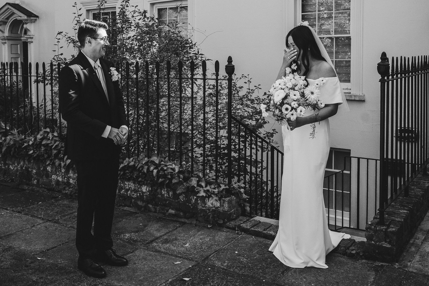 Black and white photo of a wedding couple standing on either side of an iron gate in front of a house exterior.