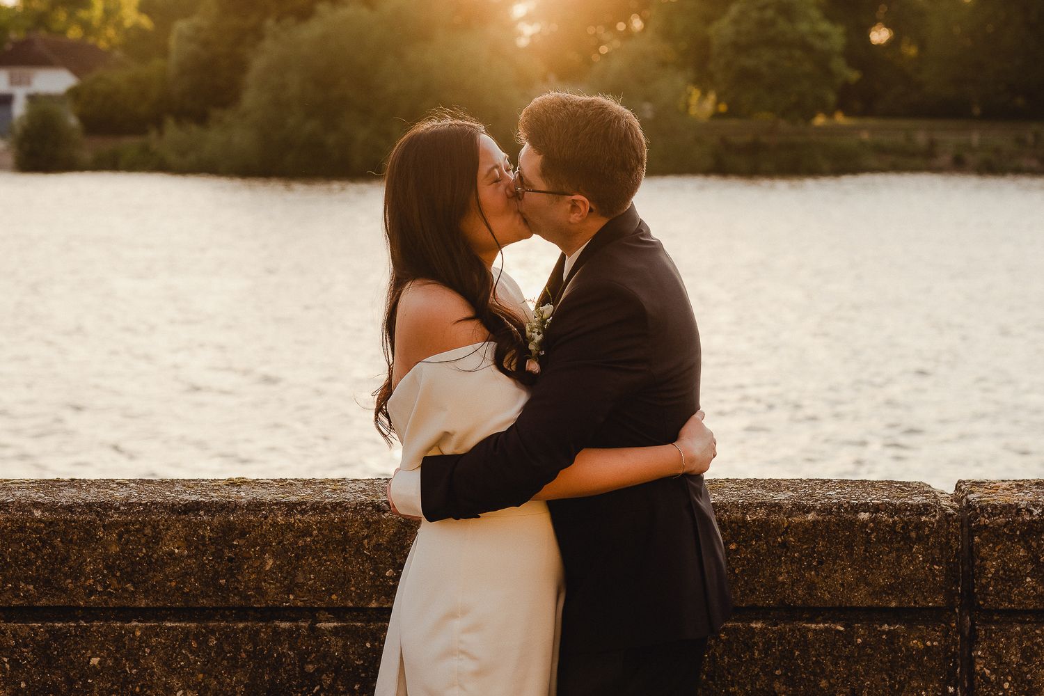 A couple in wedding attire share an intimate kiss at sunset by a stone wall overlooking a lake.