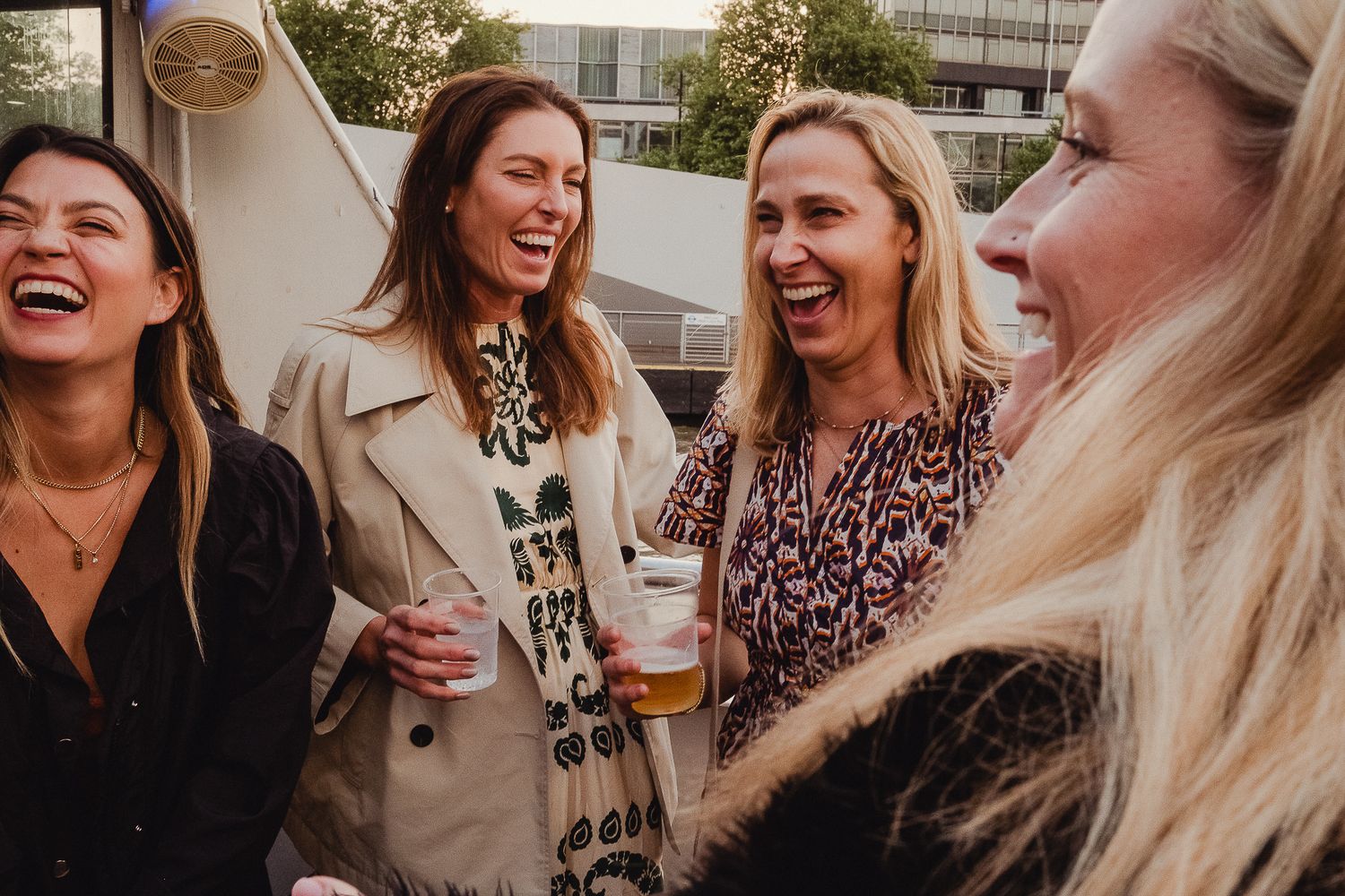 A group of friends laughing together outdoors while enjoying beverages and dressed in casual attire.