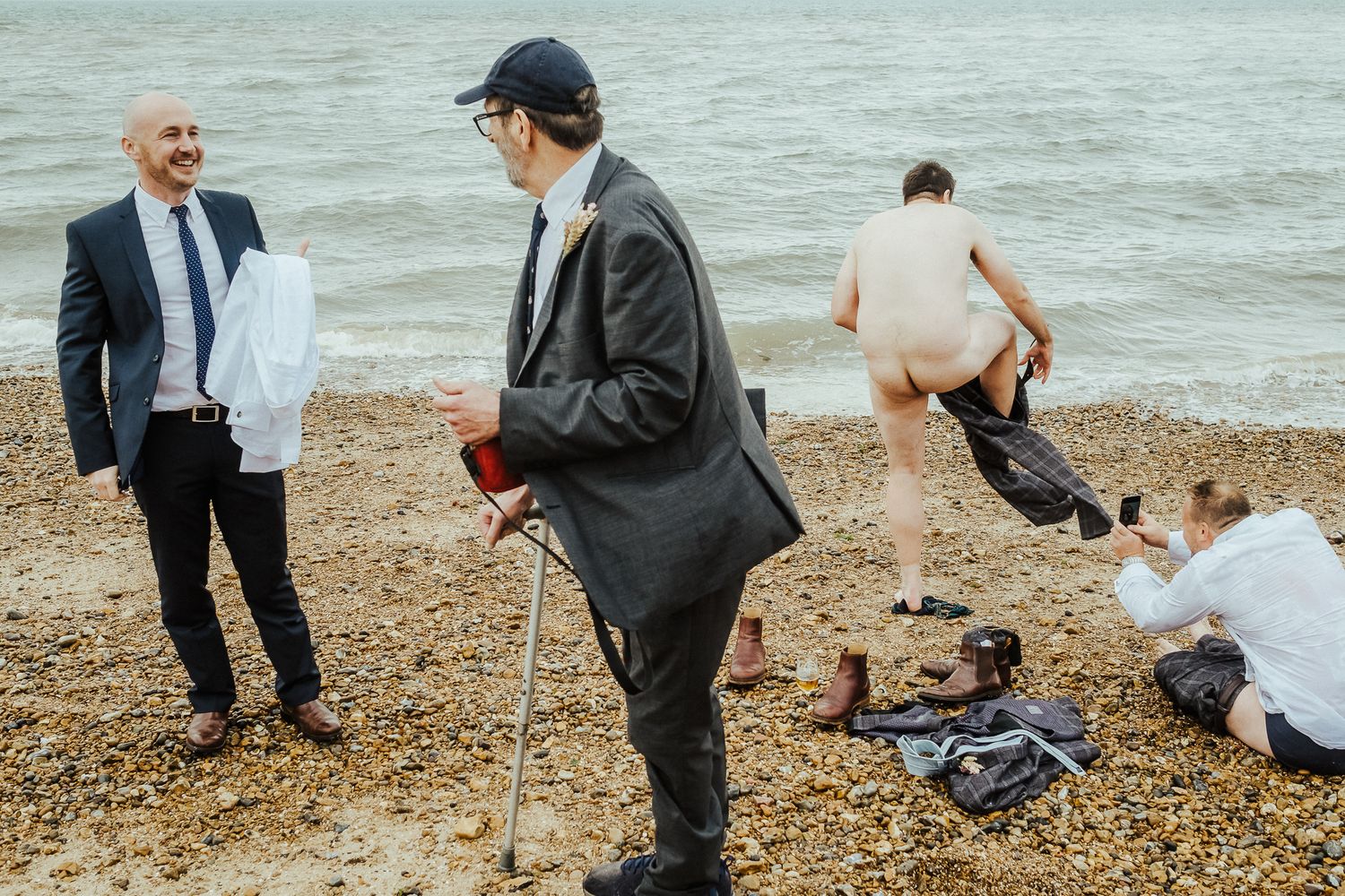 Wedding guests in formal attire gather on beach while someone swims in background.