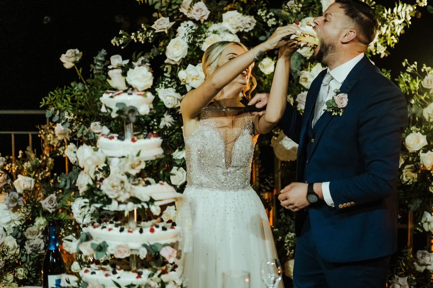 Wedding couple sharing champagne under an elegant floral arch with white roses.