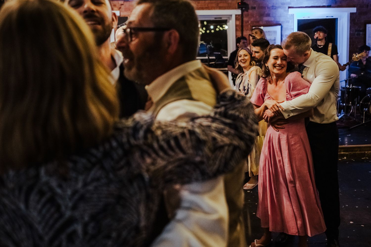 Couples slow dancing at a wedding reception with pink-dressed guests in the background.