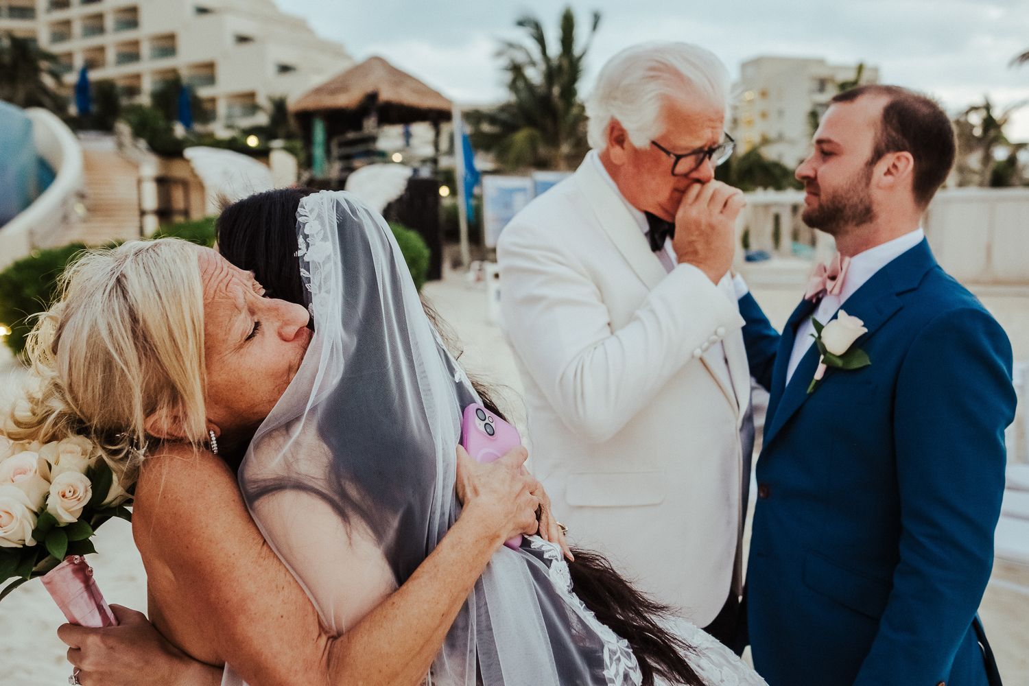 Emotional wedding photo of family members embracing while elderly man in white suit reacts.