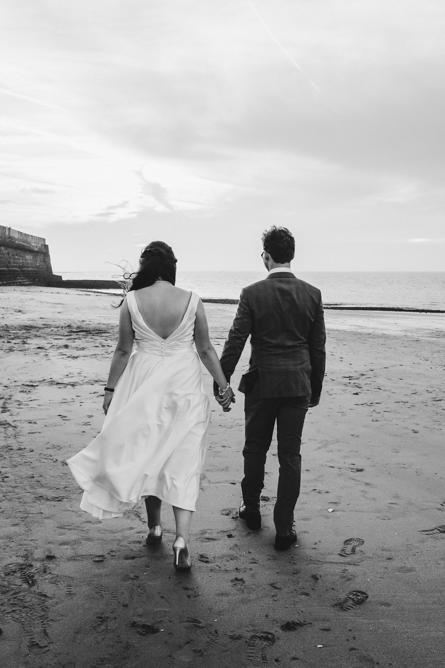 Black and white photo of a couple walking hand in hand on a beach at low tide.