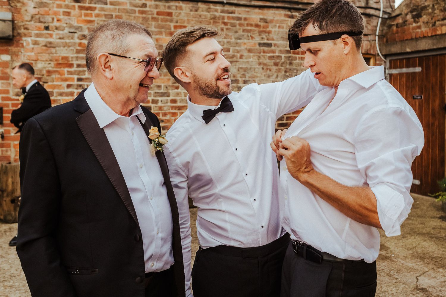 Three men in formal attire adjusting their bowties against a brick wall.