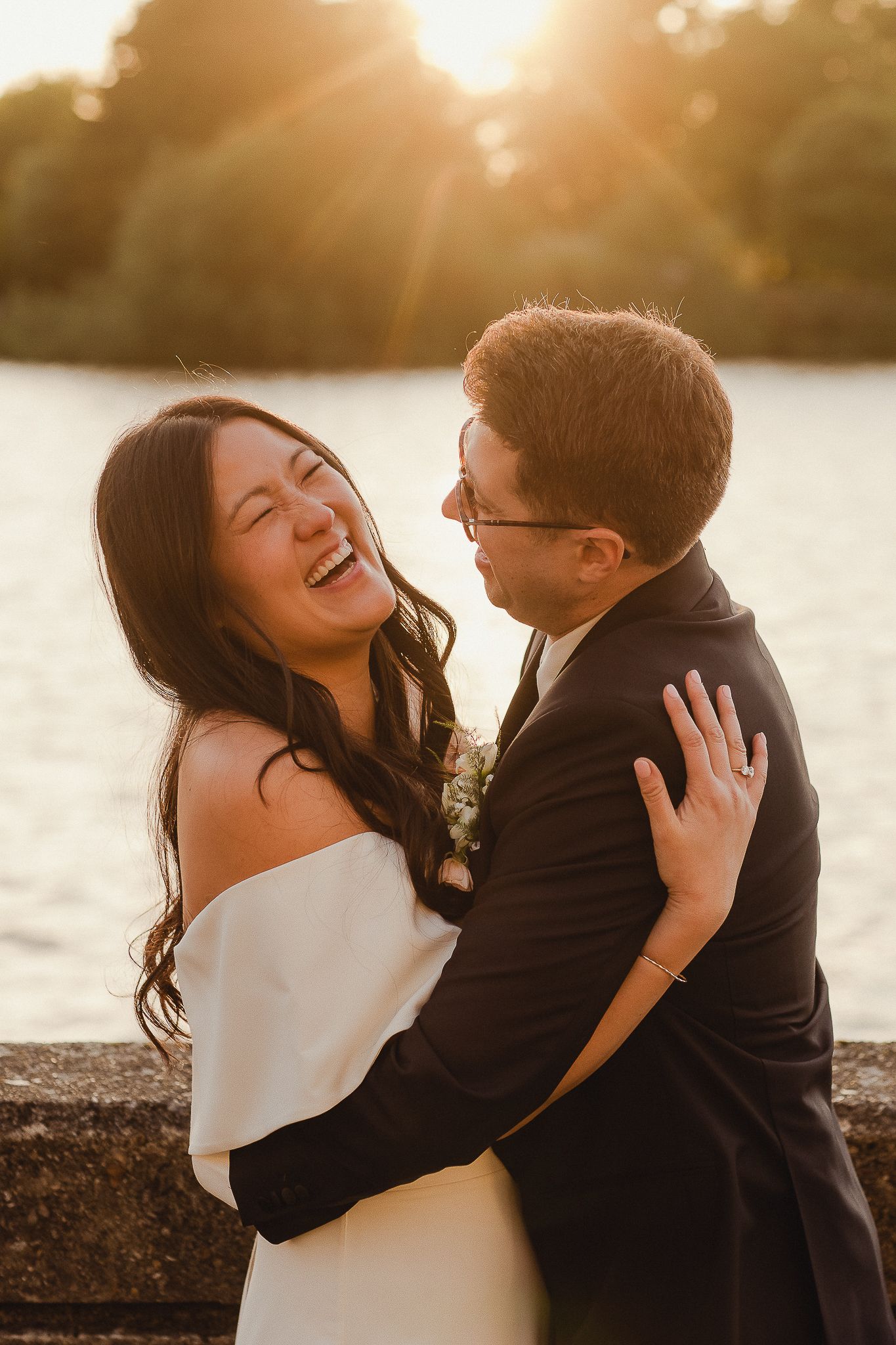 Happy couple embraces and laughs together during golden hour sunset by the water in formal wedding attire.