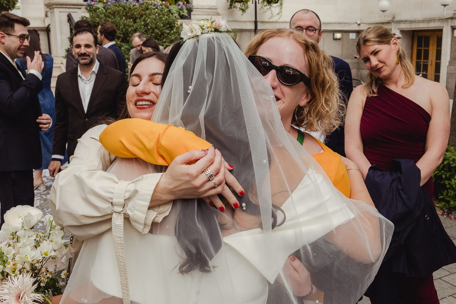 A person in a white wedding dress with a veil embraces someone in a yellow sweater while guests look on at a celebration.