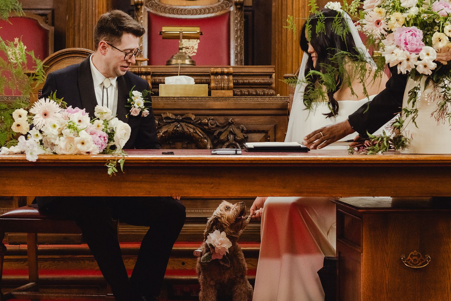 Two people sit at a wooden altar with floral arrangements during an indoor wedding ceremony.