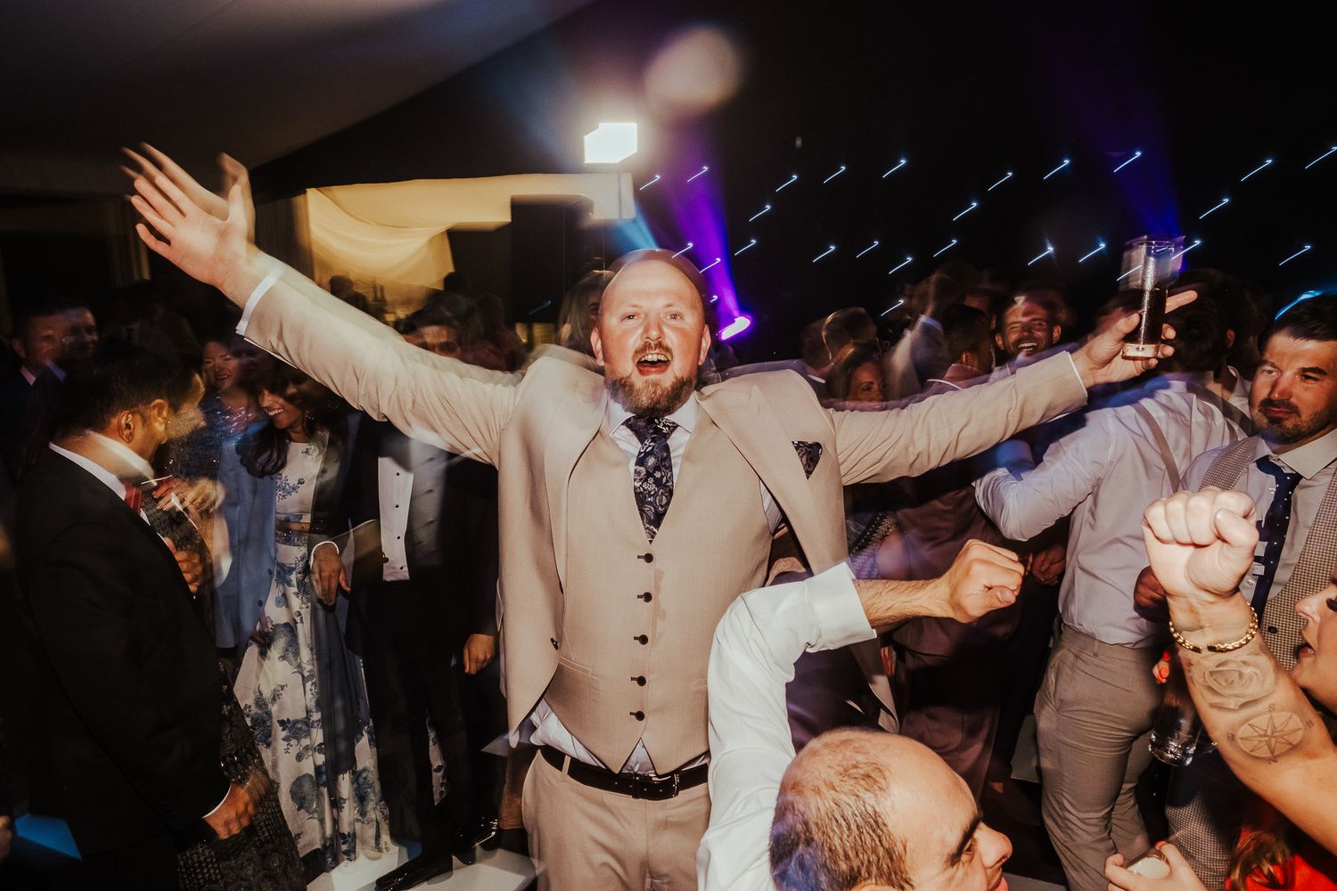 A joyful wedding guest in a light colored vest celebrates with arms raised on a lively dance floor at night.