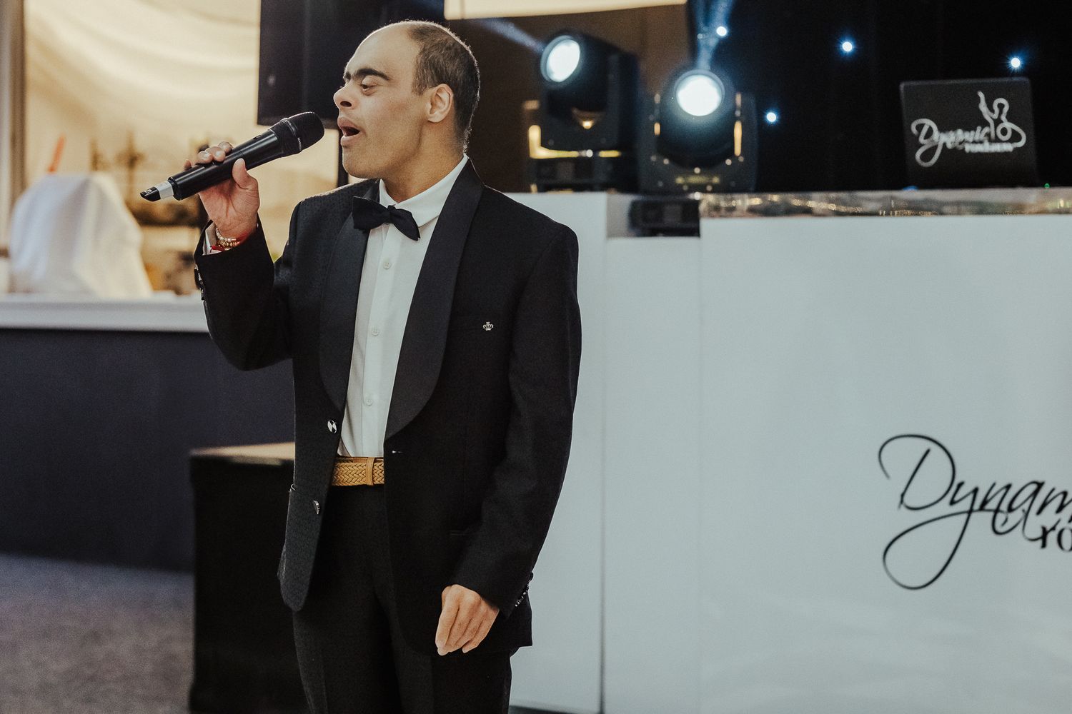 A performer in a black tuxedo sings into a microphone at an evening event next to a branded display.