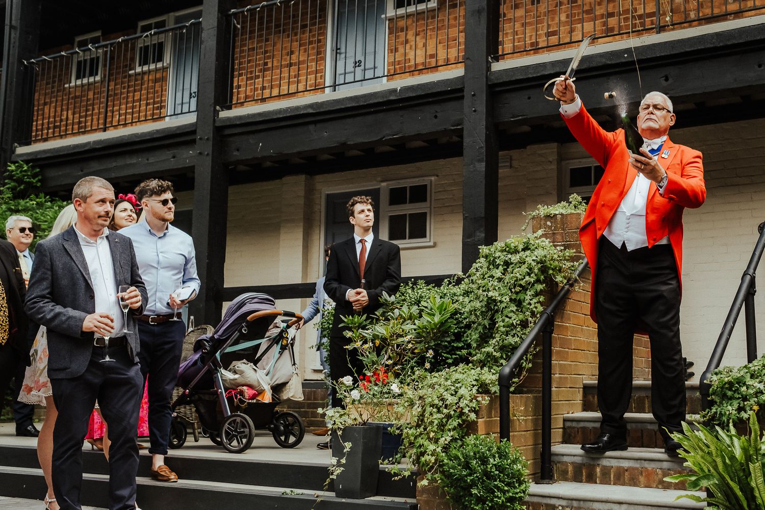 Wedding guests gather outside a brick building while someone in orange makes an enthusiastic gesture.