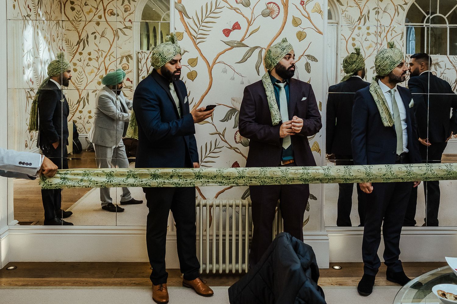 Several men in formal attire stand near a decorative wall with floral patterns during an event.