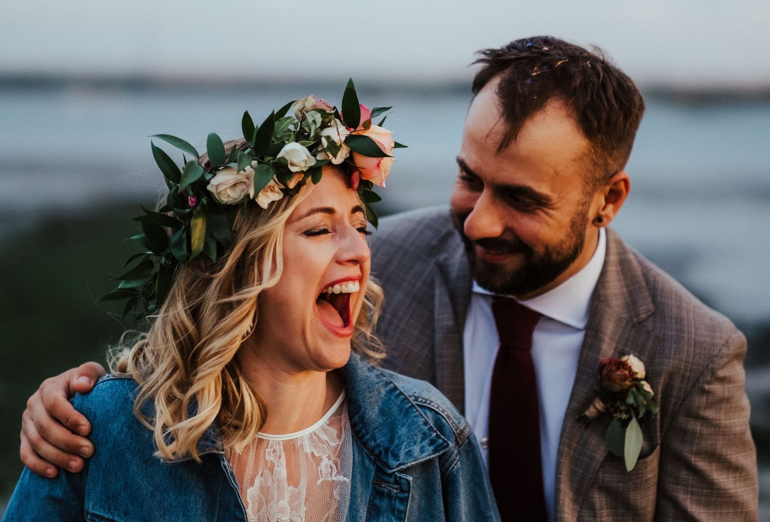 Joyful couple laughs together during outdoor wedding photoshoot with floral crown and formal attire.