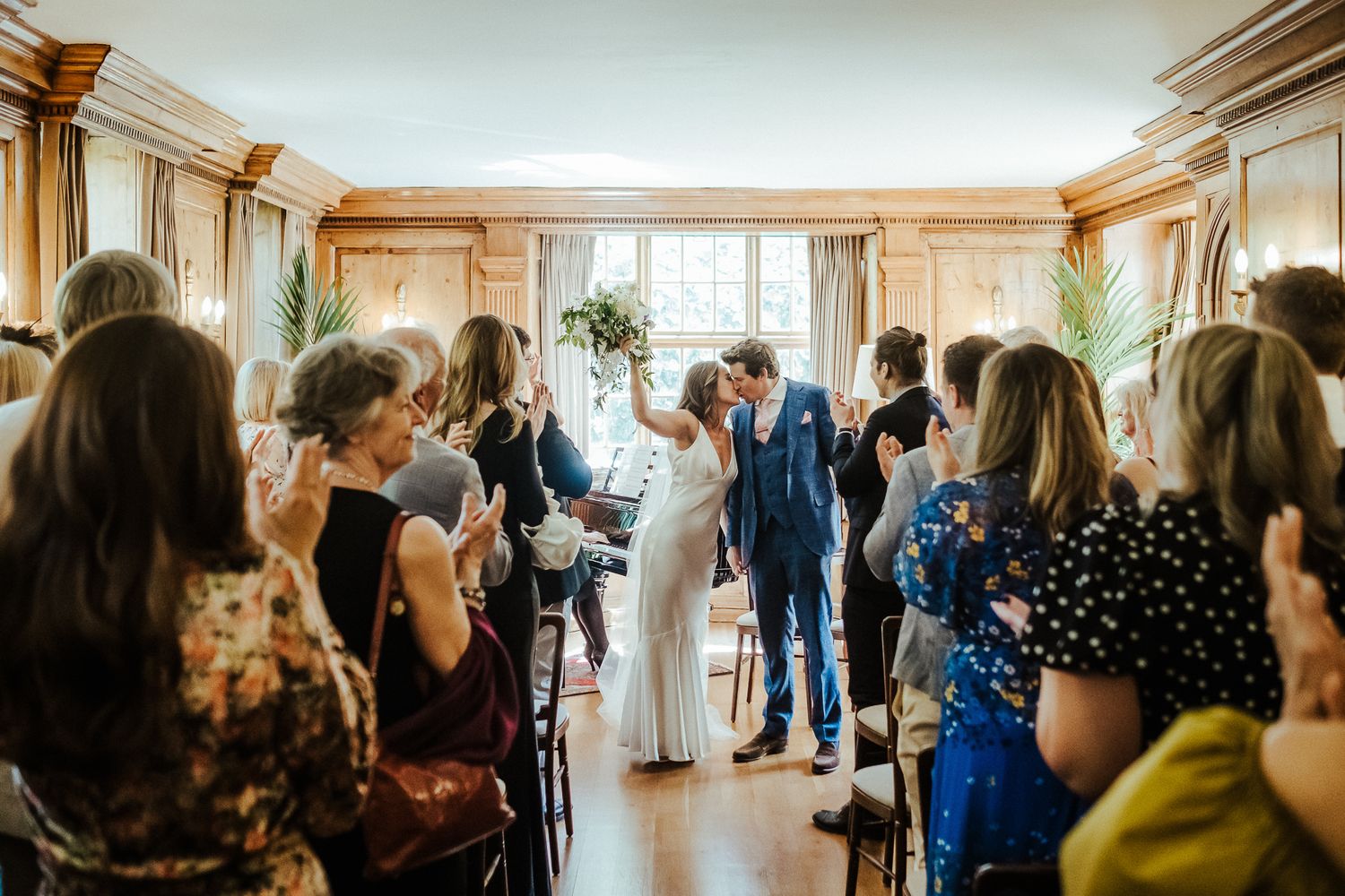Wedding guests applaud the newlyweds during their celebration in an elegant wood-paneled room.