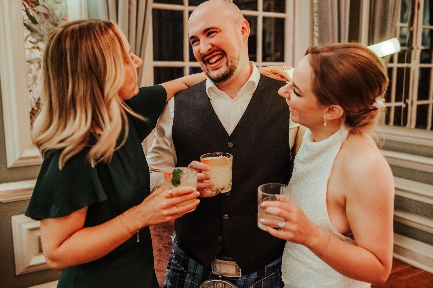 Wedding guests sharing drinks and laughter at an evening reception in warm lighting.