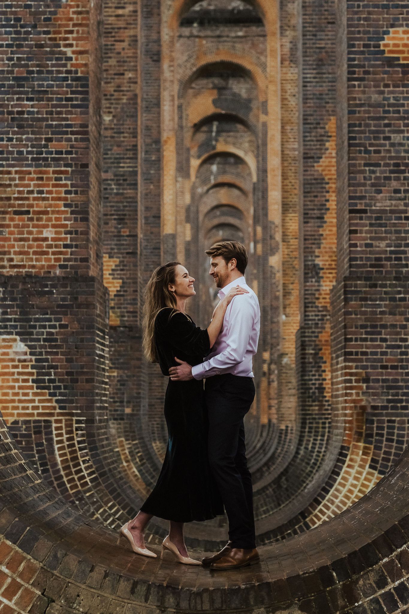 Romantic couple posing in a curved brick tunnel with repeating arches.
