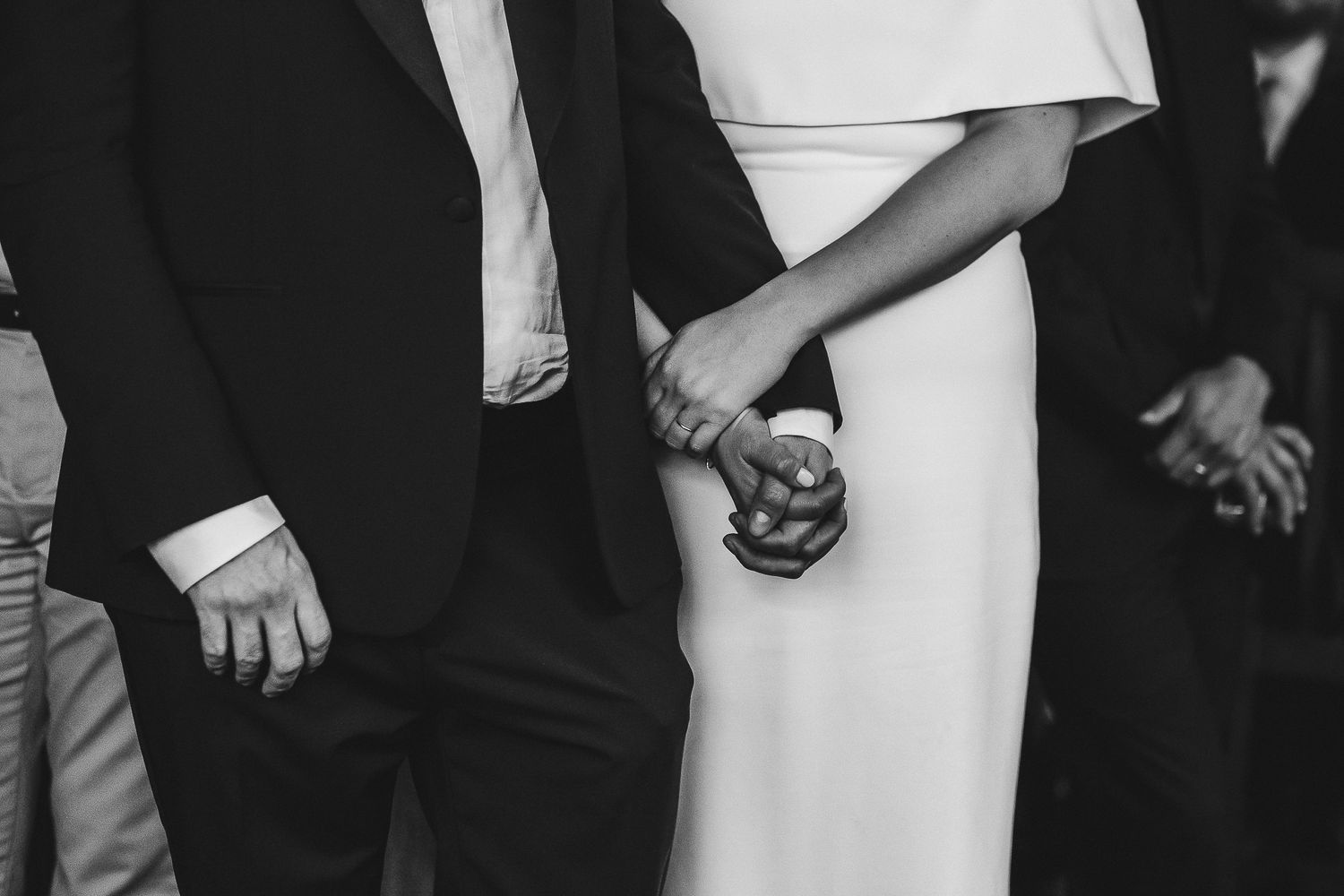 A black and white close-up photo showing a couple holding hands during what appears to be a wedding ceremony.