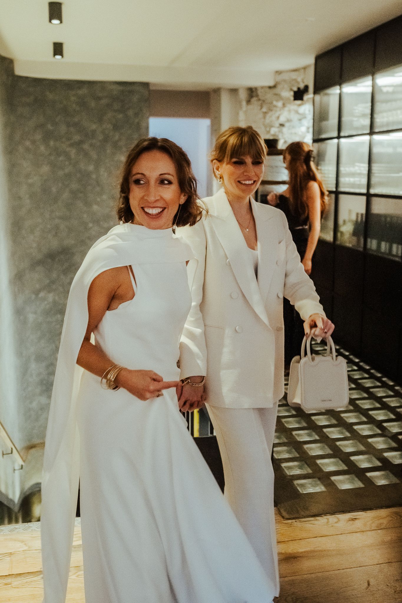 Two people in elegant white outfits share a joyful moment in a modern interior setting.