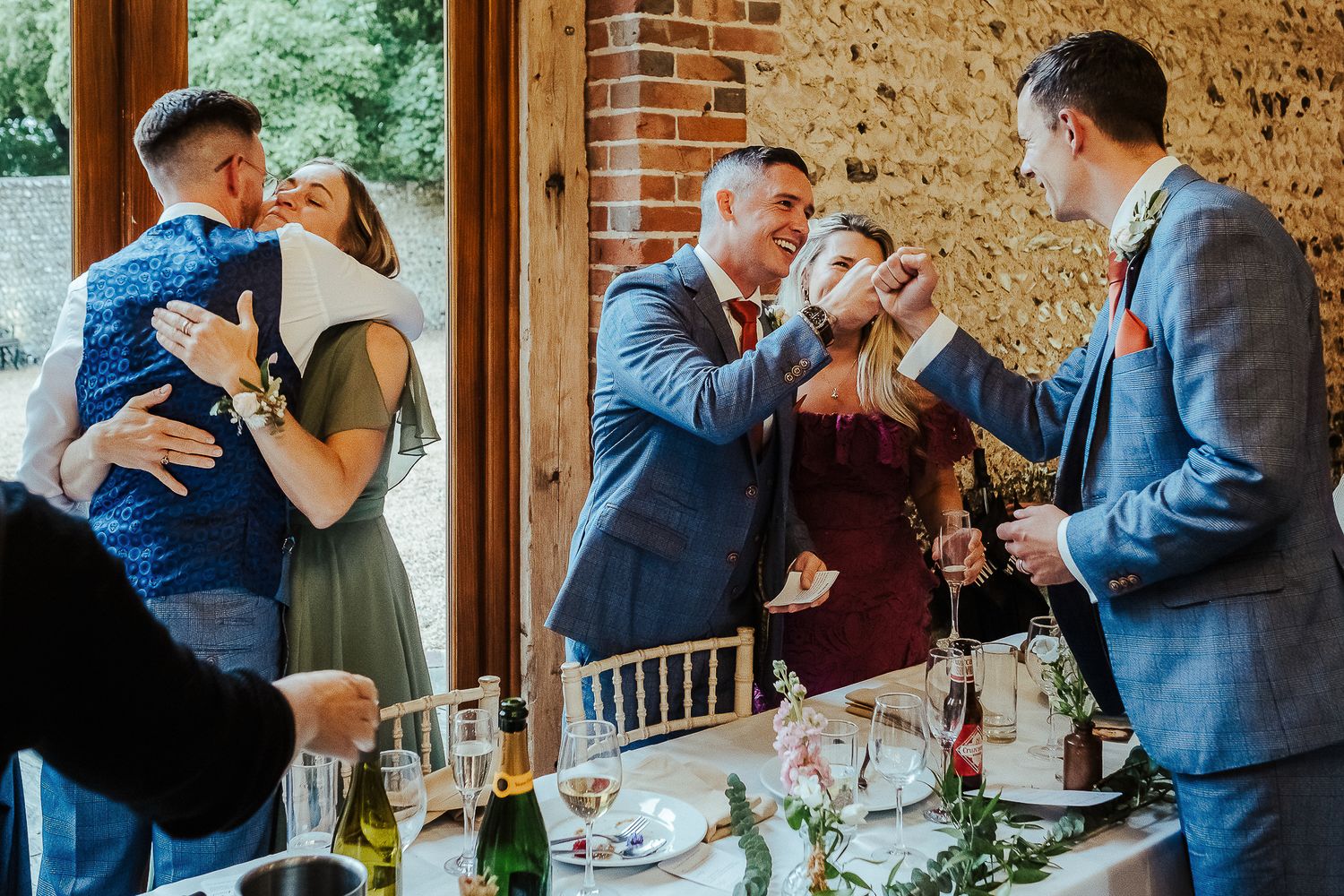 Wedding guests and party members celebrate with hugs in a rustic brick venue.