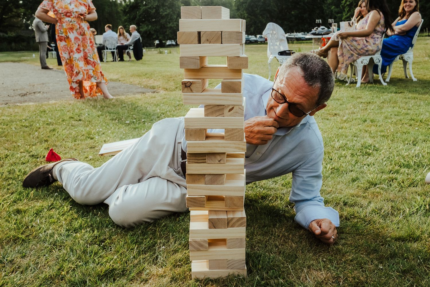 Person lying on grass carefully removing block from giant outdoor Jenga tower at wedding.