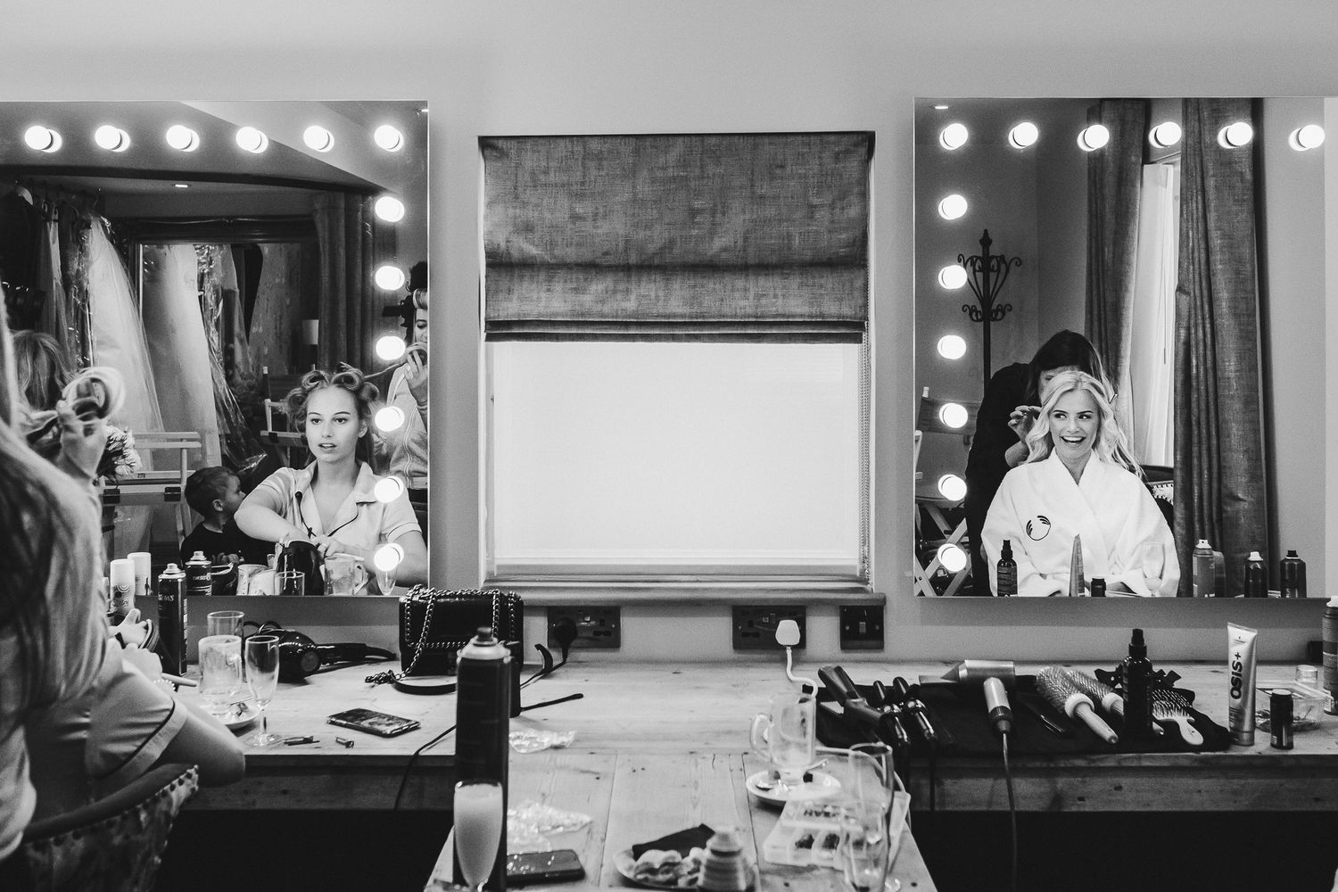 Black and white image of wedding preparations in a dressing room with illuminated vanity mirrors.