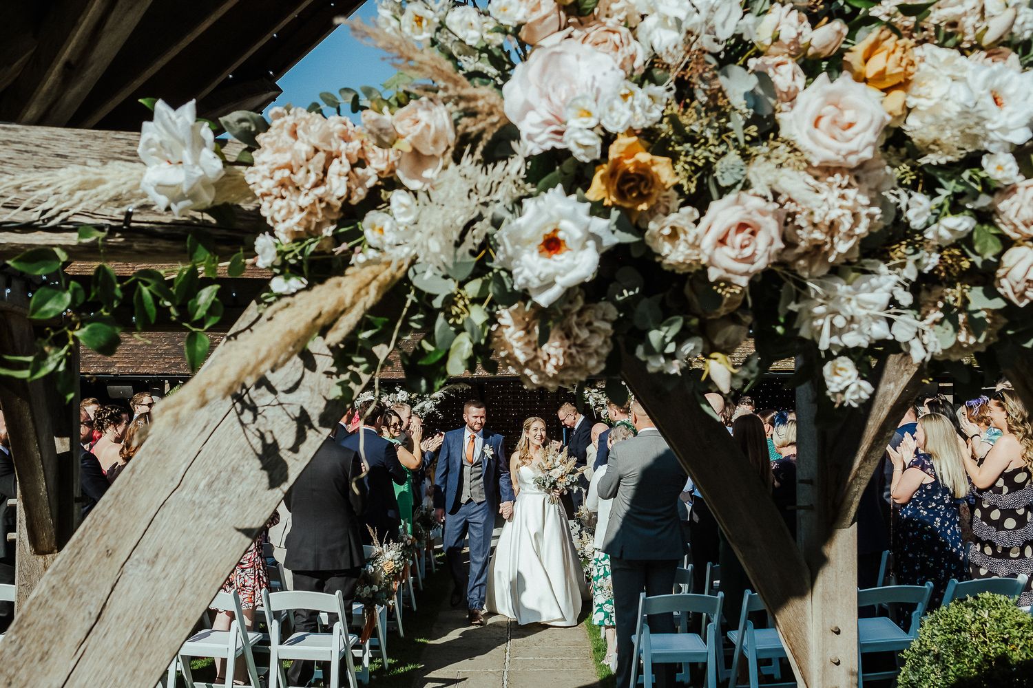 Wedding ceremony under floral arch with white chairs and guests.