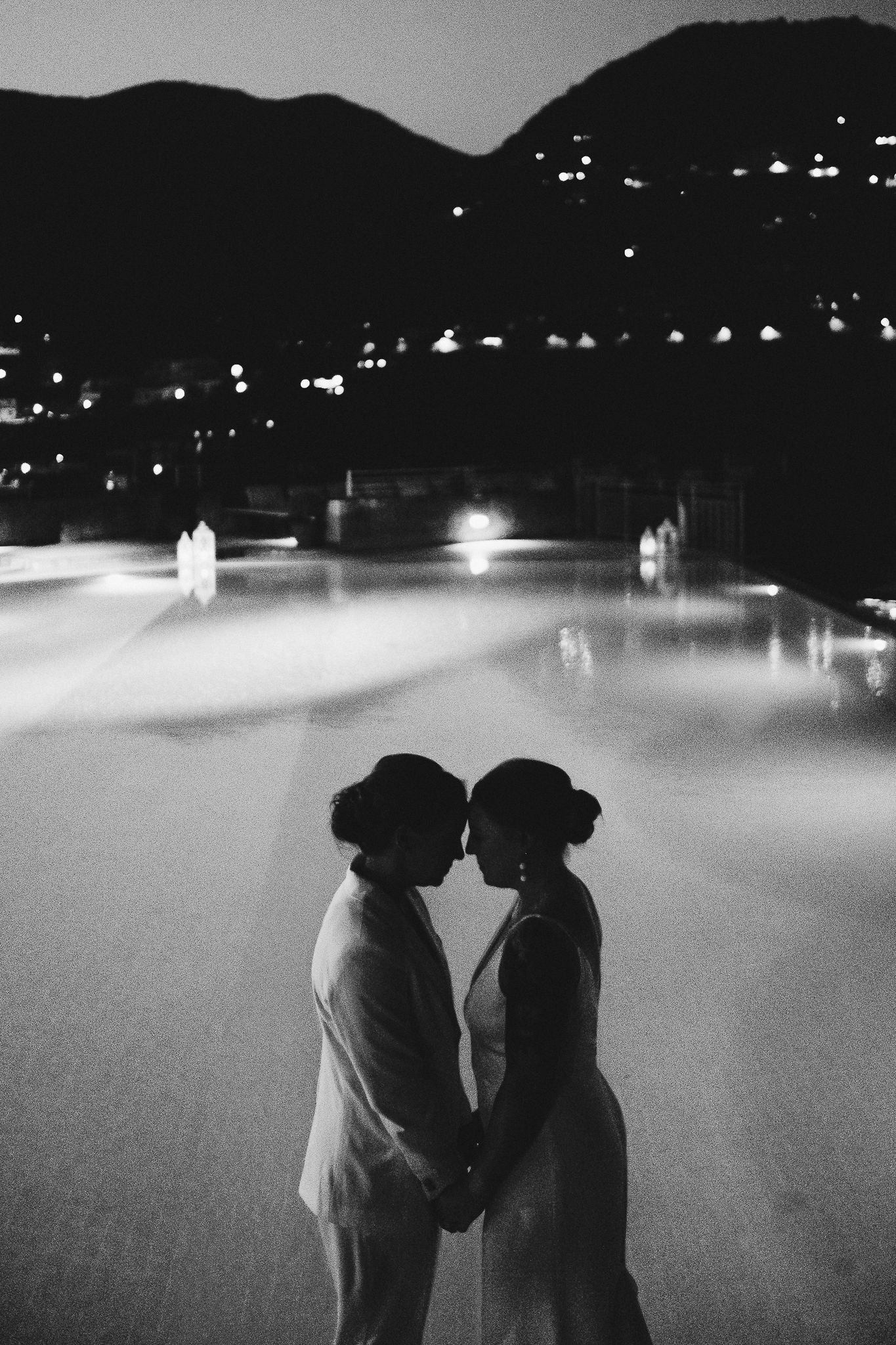 Two silhouettes share an intimate moment on an illuminated ice rink at night with mountain lights in the background.
