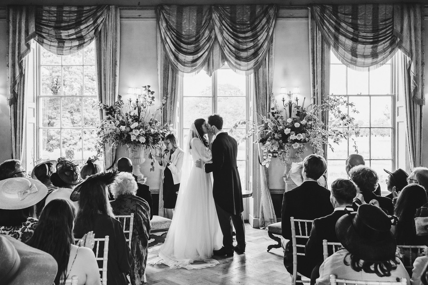 A black and white wedding ceremony in an elegant room with draped windows and floral arrangements.