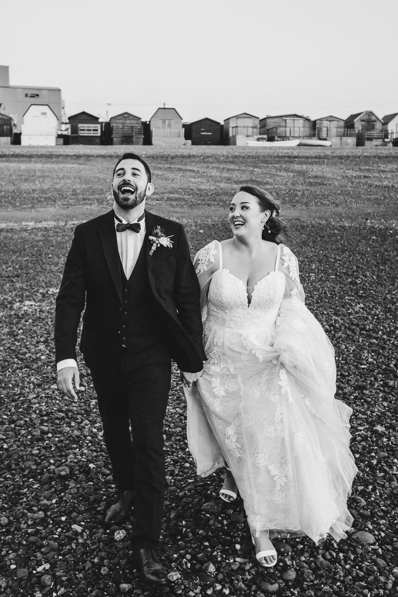 Joyful couple laughs while walking on beach with beach huts in background.