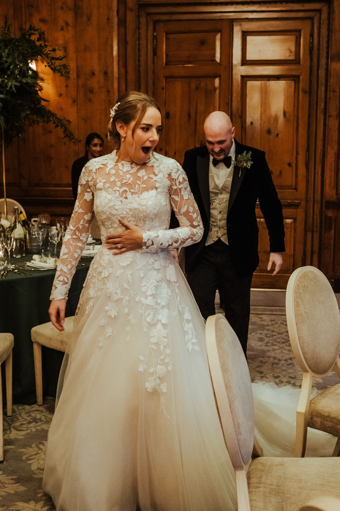 Bride in long-sleeved lace gown shares a candid moment with guest in formal wedding reception room.