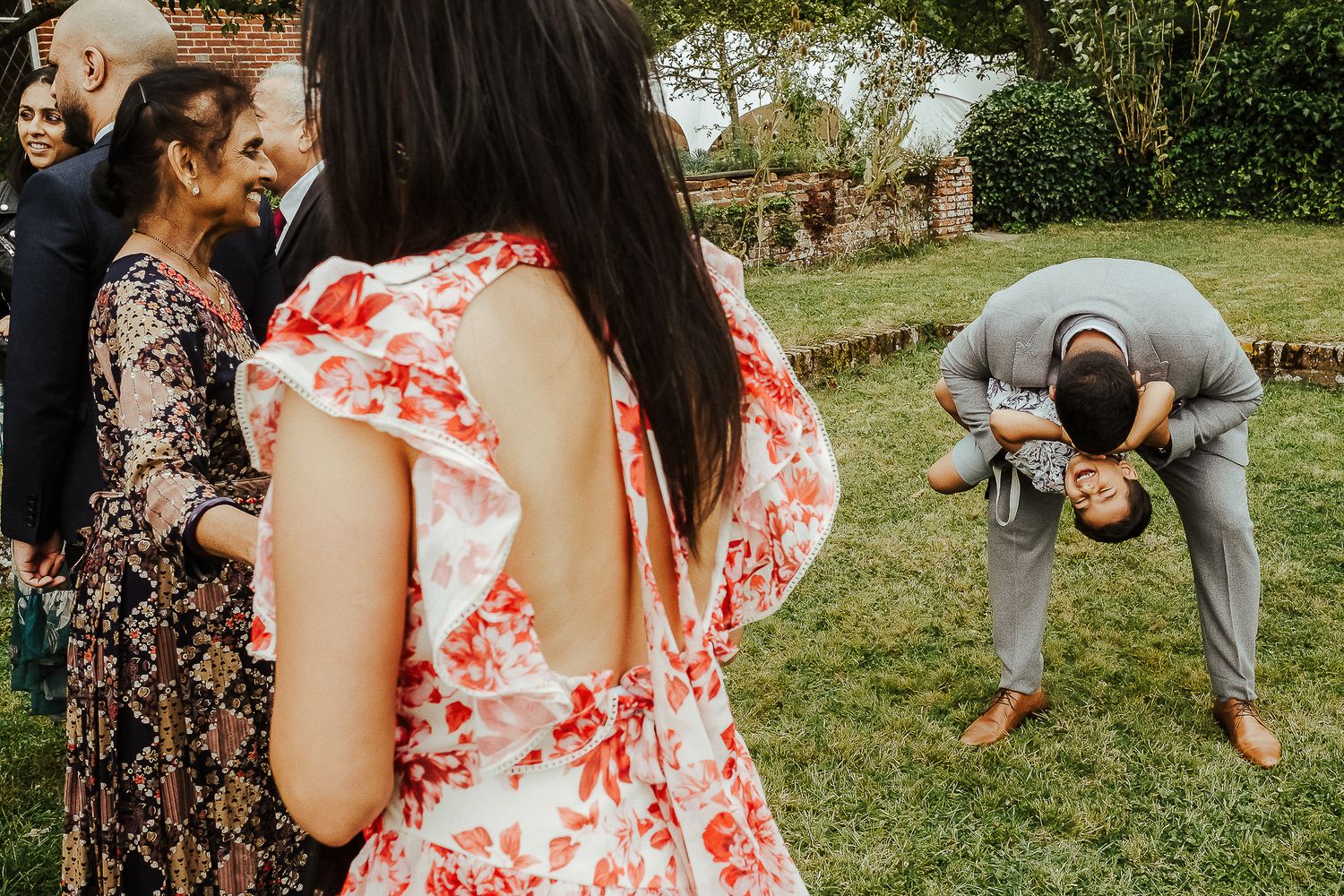 Wedding guests in floral dresses gather outdoors while someone bends down in laughter.