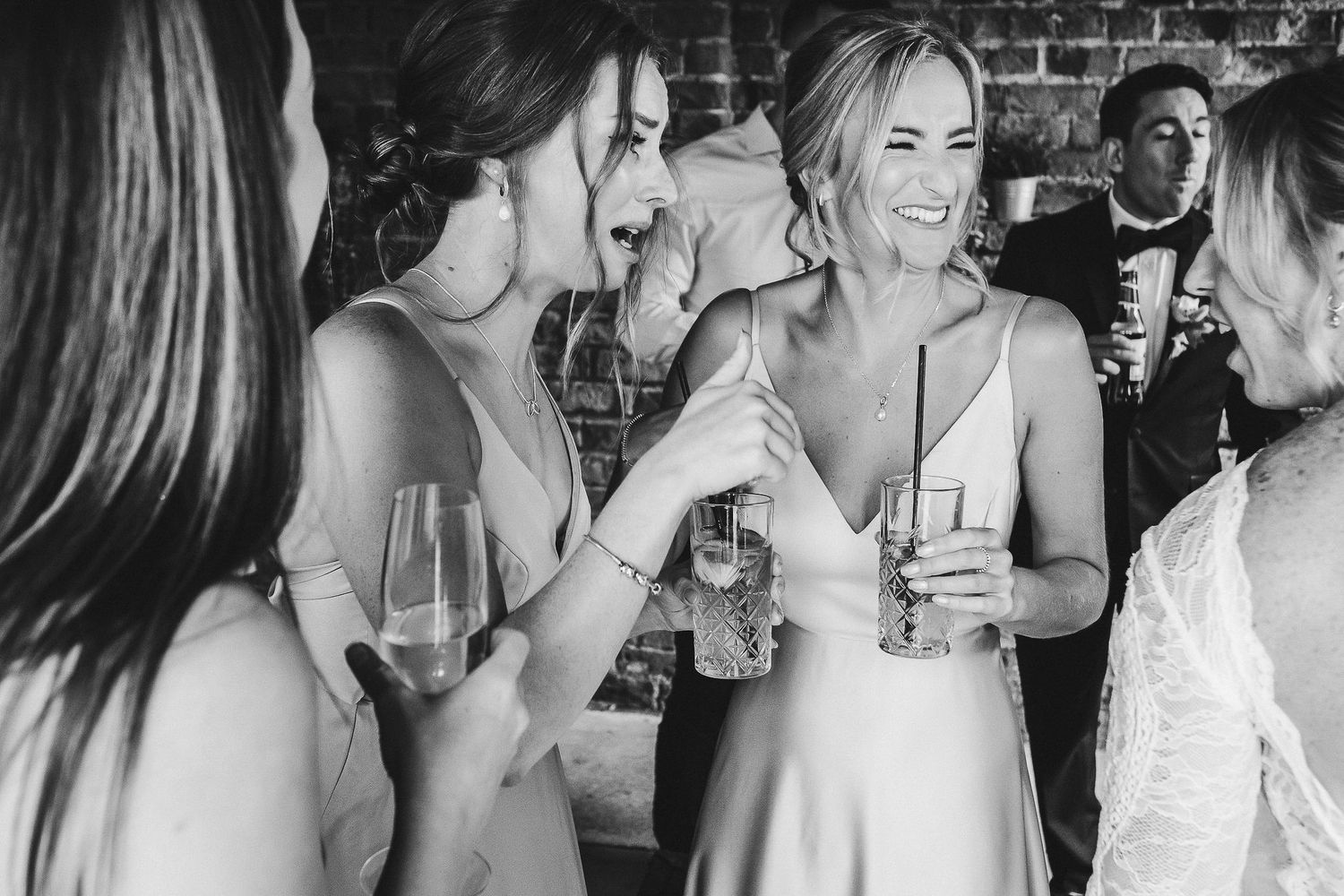 Group of wedding guests in formal attire share drinks and laughter at brick-walled reception venue.