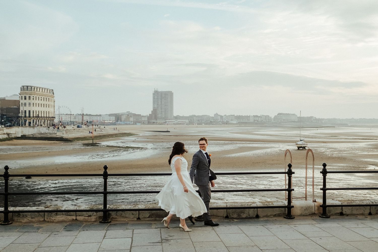 Couple in wedding attire standing by seaside railings with coastal views.