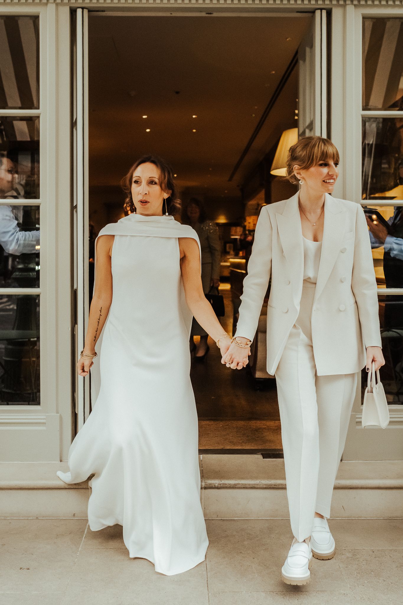 A couple in elegant white outfits exiting a storefront holding hands.