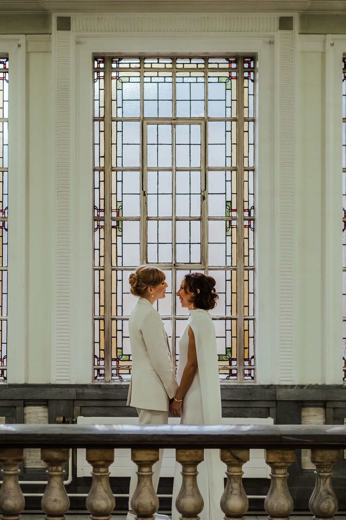 Two people in white stand together against ornate window architecture.