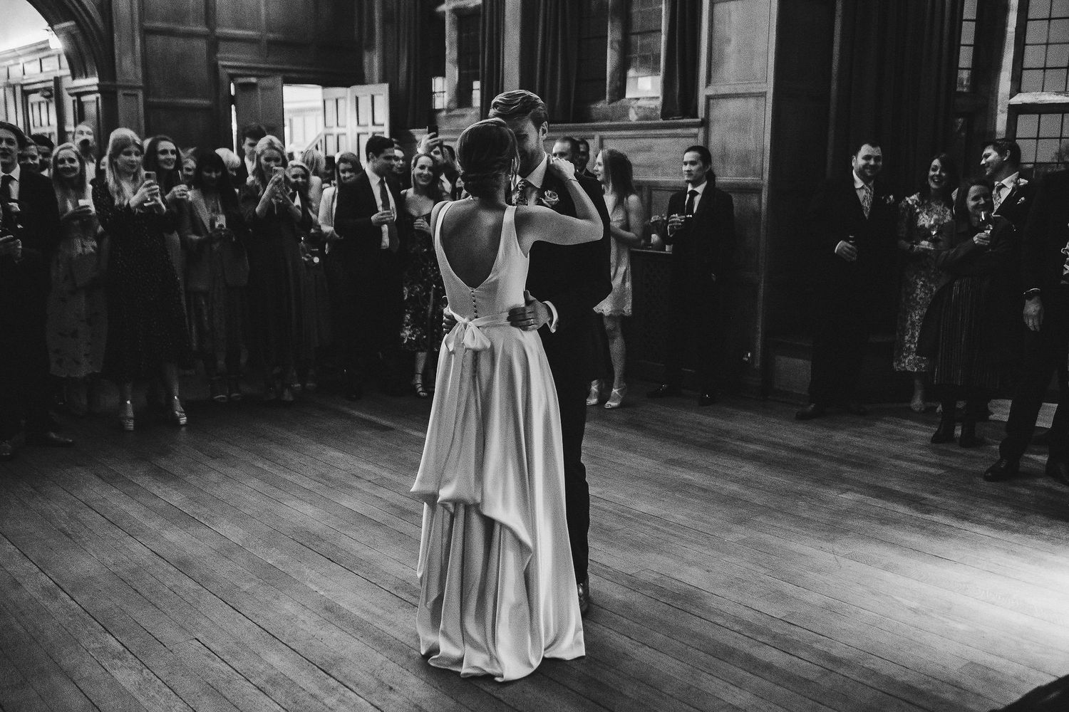 A couple shares their first dance in a grand wood-paneled ballroom surrounded by wedding guests.