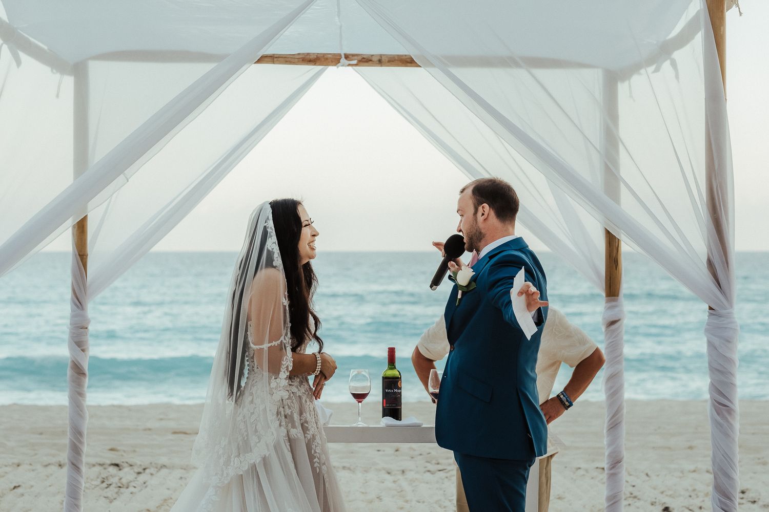 Couple exchanging vows under a white beach canopy with ocean backdrop.