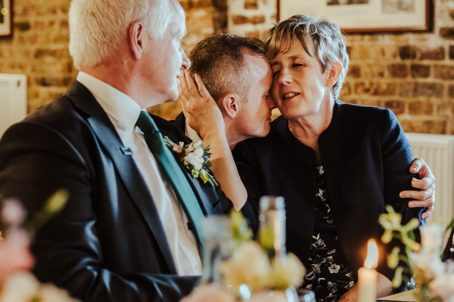 Intimate candid moment shared between wedding guests during reception at brick-walled venue.