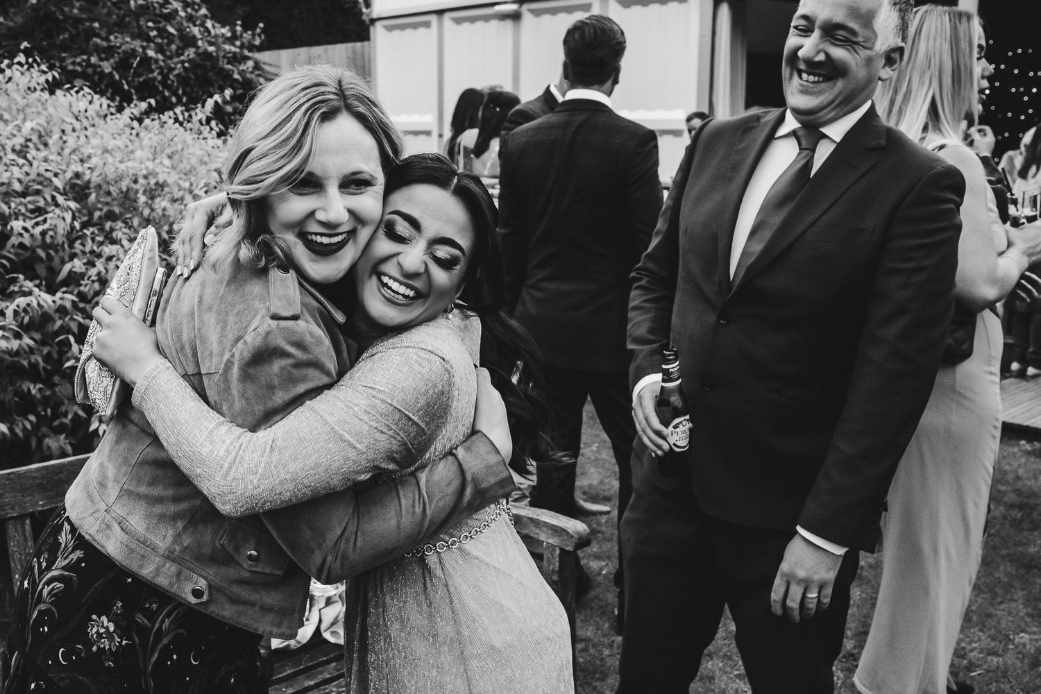 Black and white candid photo of two people sharing a happy embrace at an outdoor celebration while others look on.