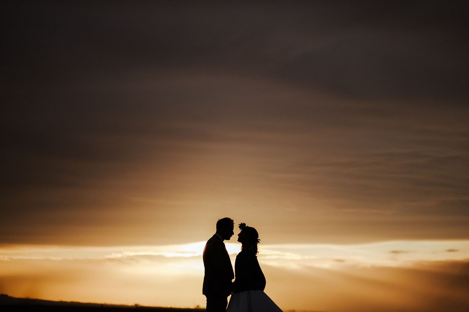 Silhouetted couple shares romantic moment against dramatic sunset sky at coastal wedding ceremony.