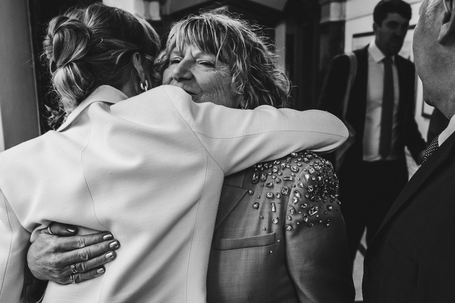 Black and white photo of emotional wedding embrace with guests in formal attire.