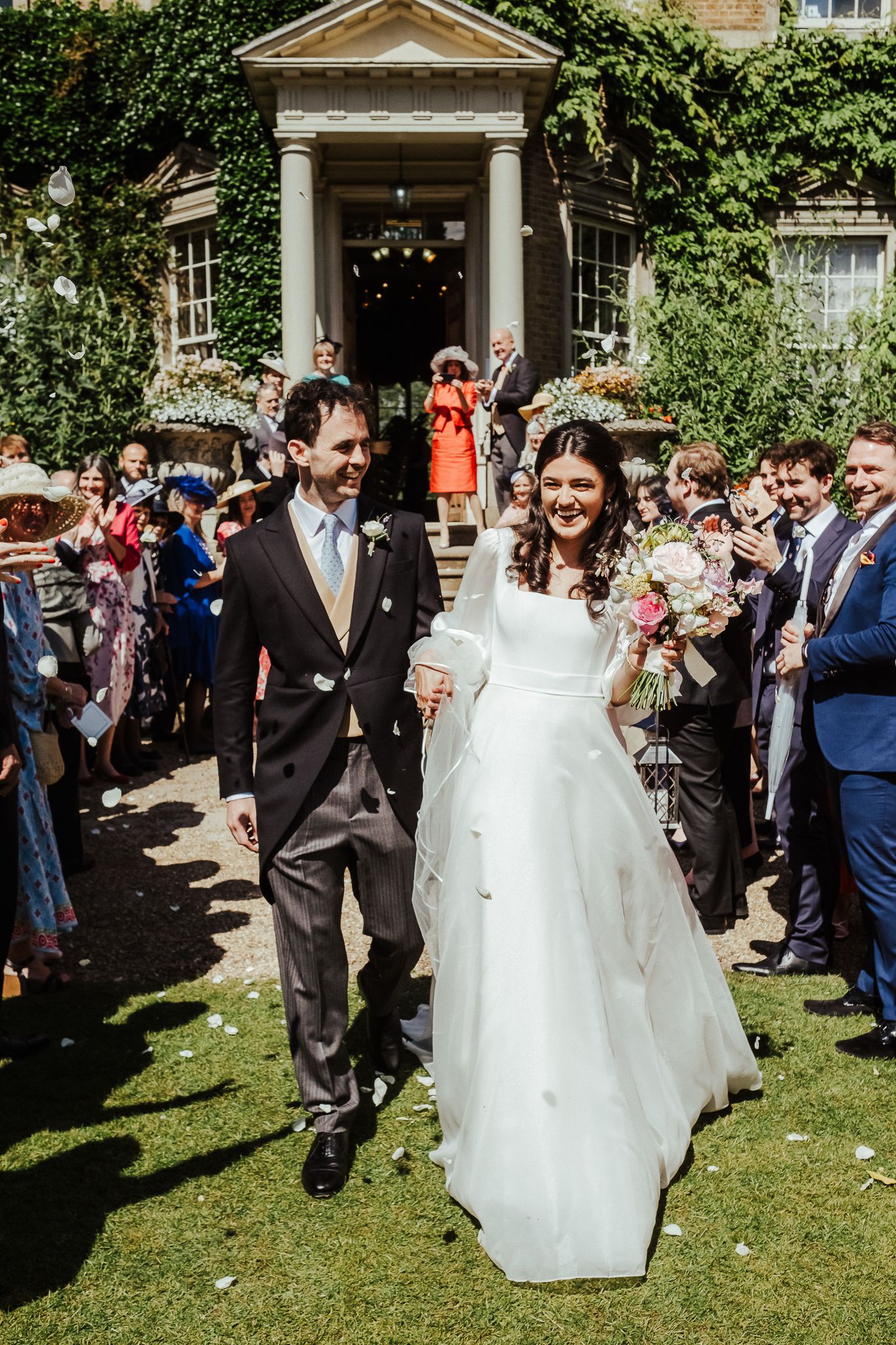 Newlyweds walking down garden path while guests throw confetti in celebration.