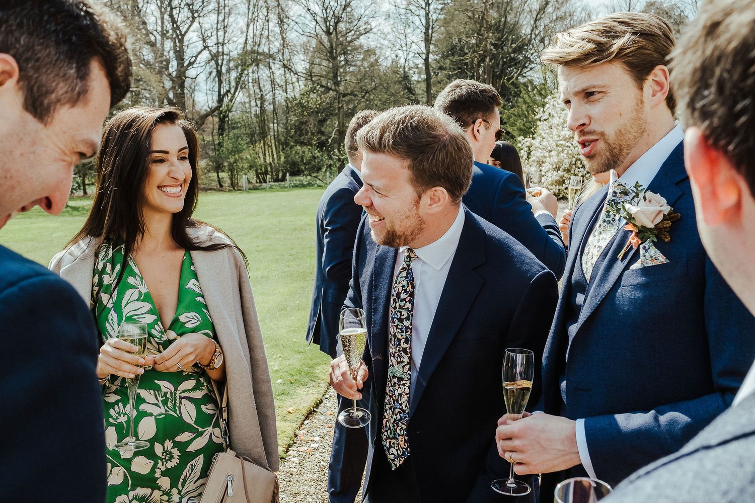 Wedding guests in formal attire socialize and enjoy drinks in a garden setting.