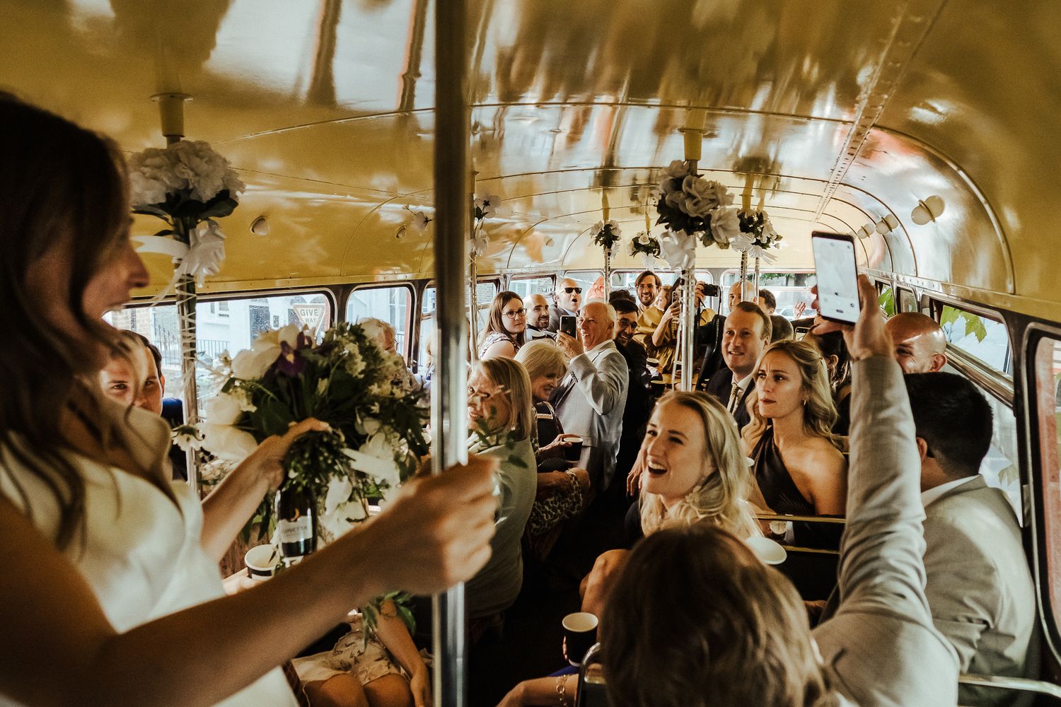 Wedding party celebrates inside a decorated vintage bus with raised hands and flowers.