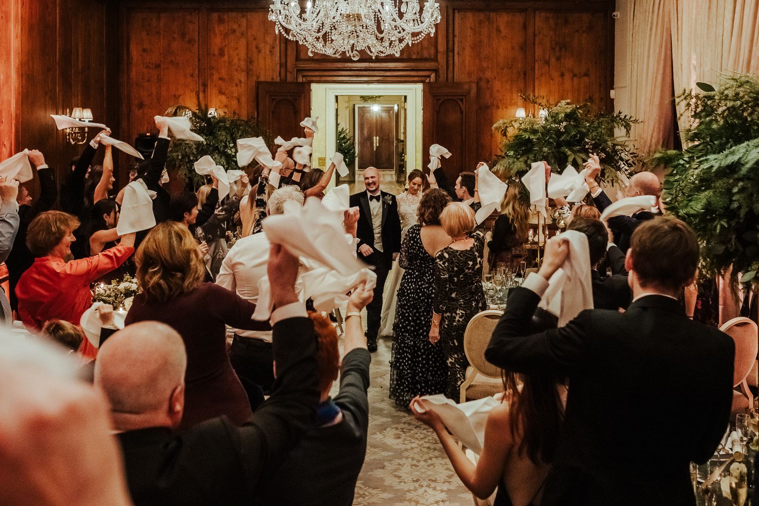 Lively wedding reception scene with dancing guests in ornate wood-paneled room with chandelier.