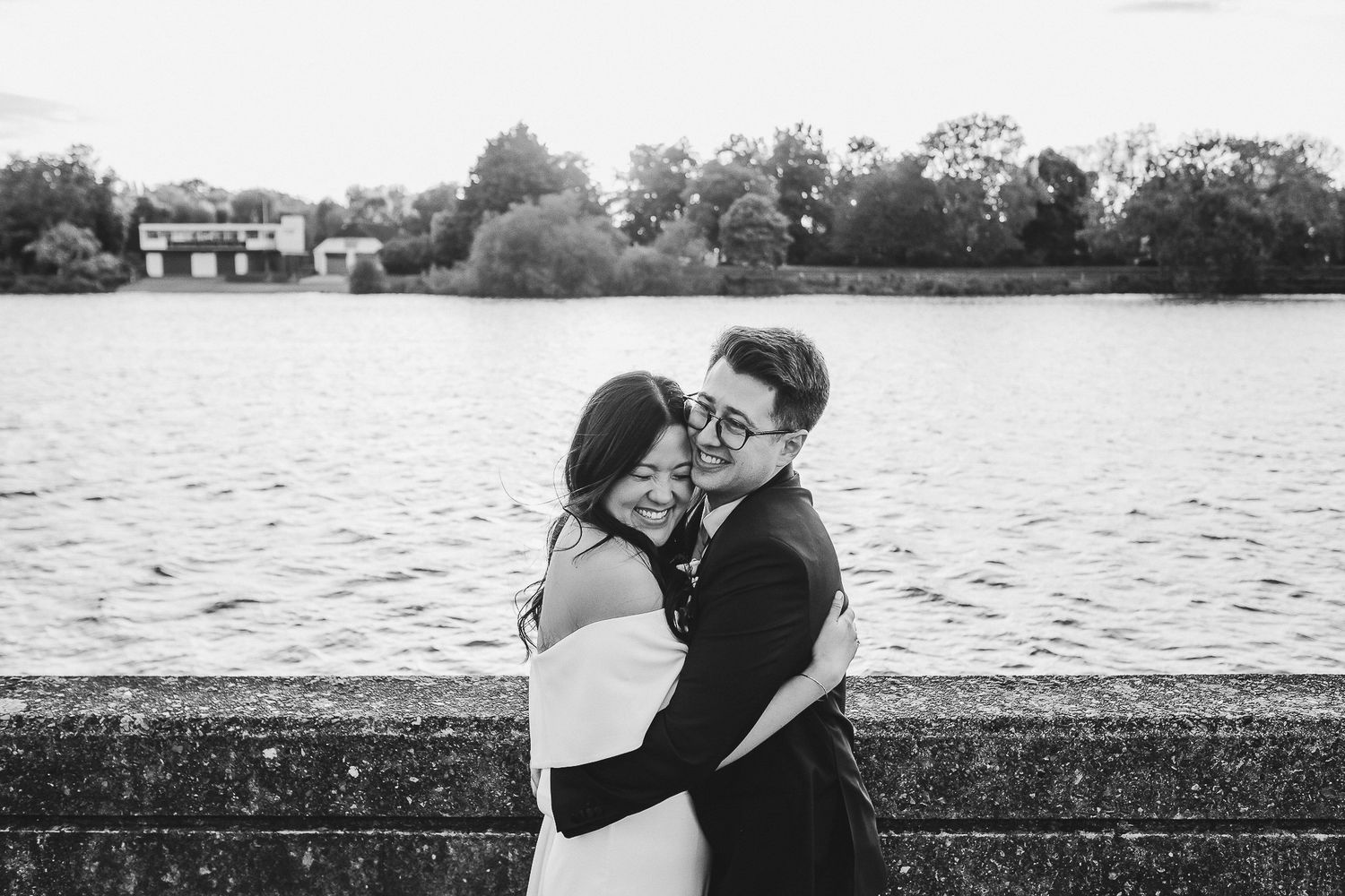 A couple embraces lovingly by a lake with trees in the background in black and white.