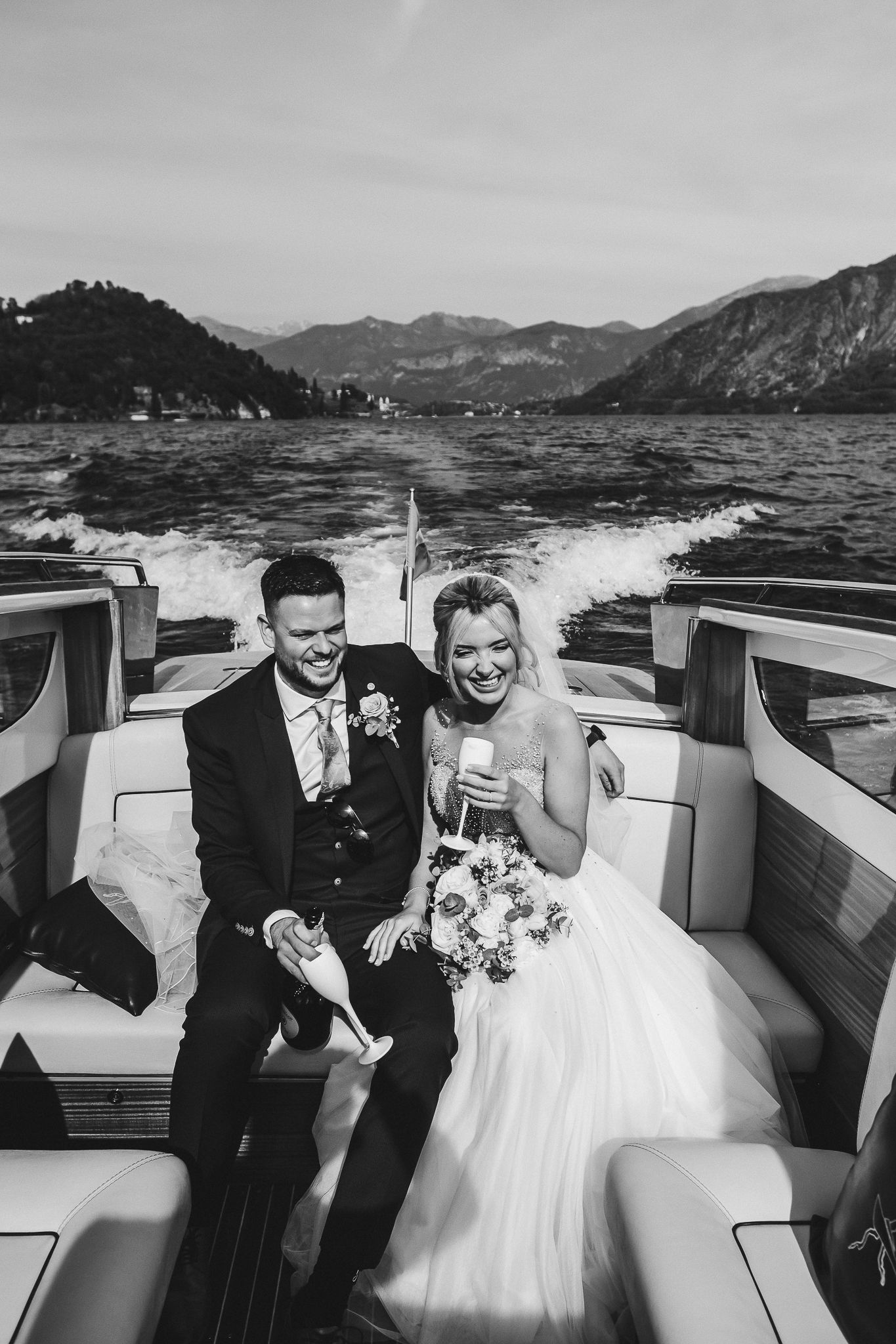 Black and white image of newlyweds laughing during boat ride with wake trailing behind.