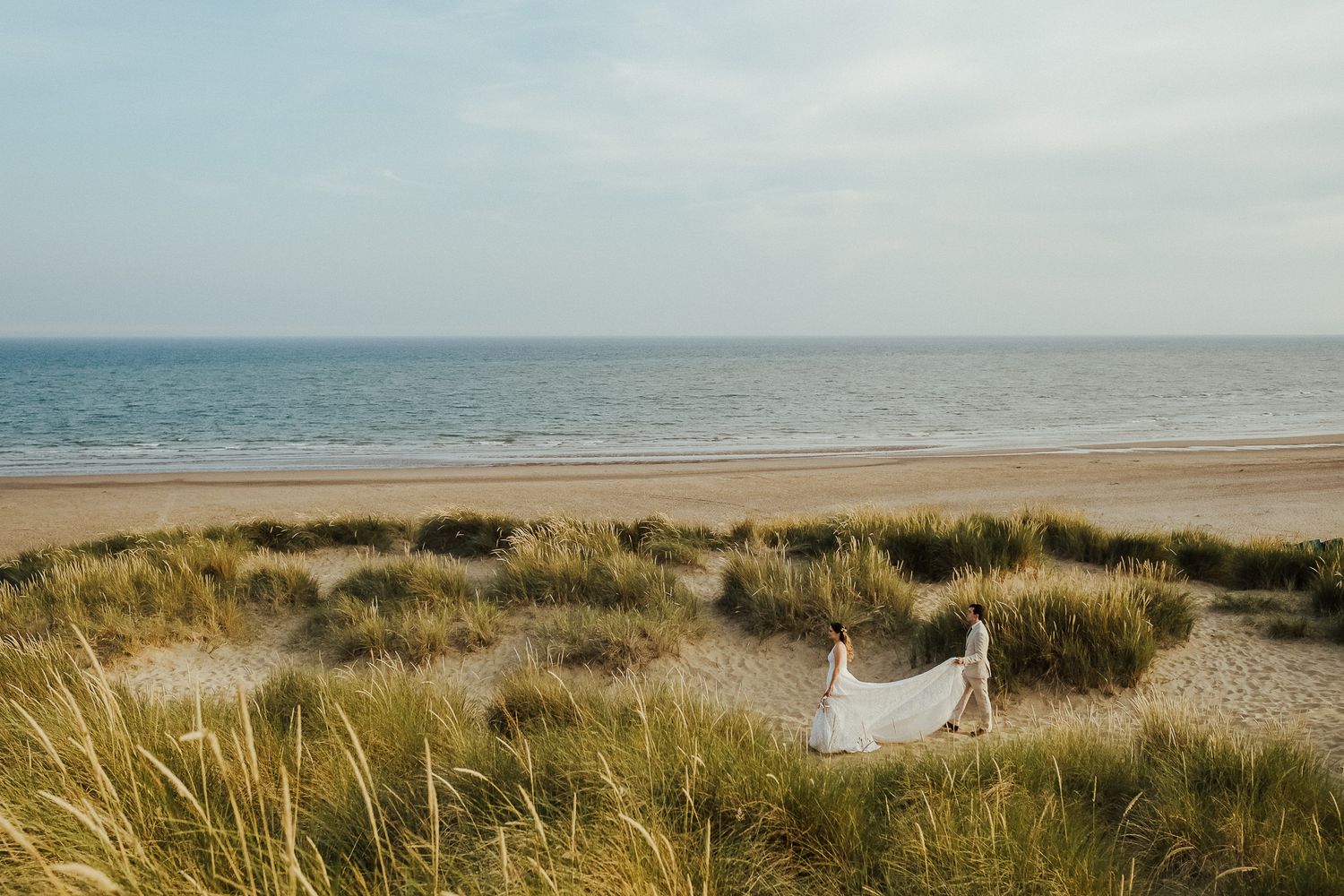 A wedding dress lays on sand dunes with ocean and beach horizon in the background.