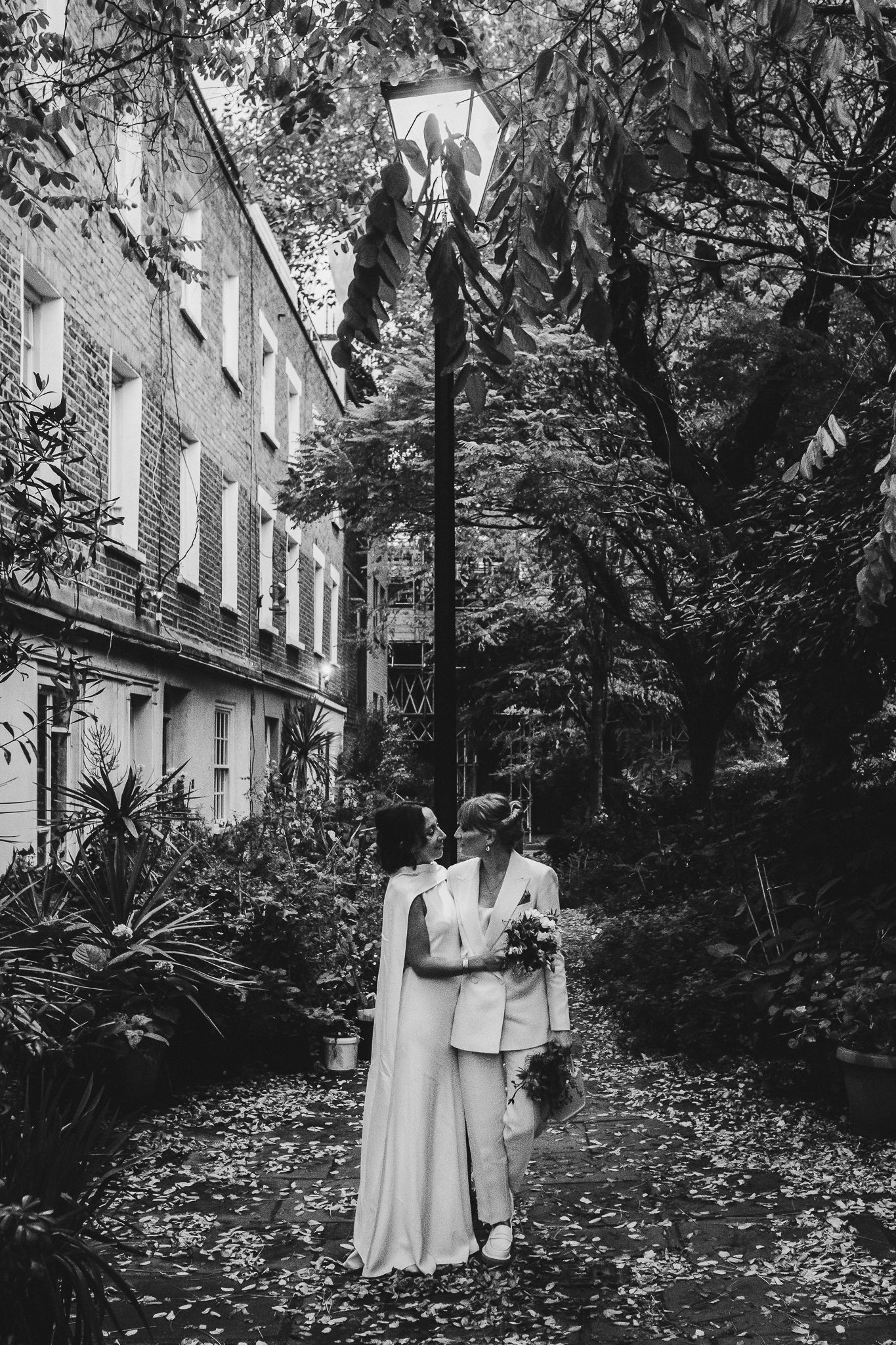 Black and white photo of couple in wedding attire standing together on leaf-covered garden path.